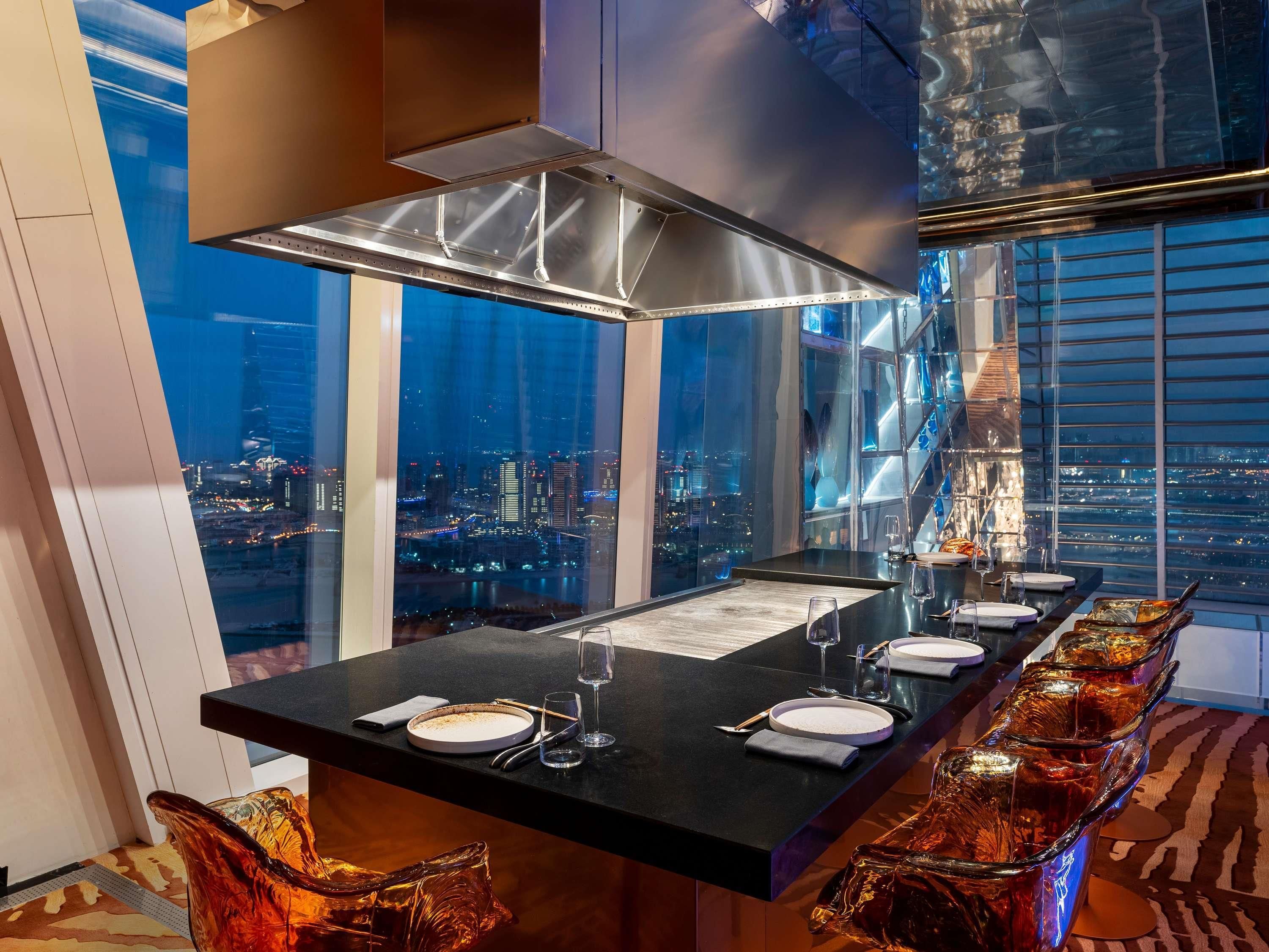 Un comedor en un edificio alto con vista a la ciudad, una cocina moderna y una larga mesa de comedor dispuesta para una comida.