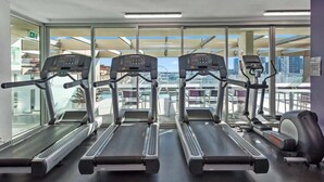 A gym with treadmills, exercise bikes, and a rowing machine, with large windows offering a city view.
