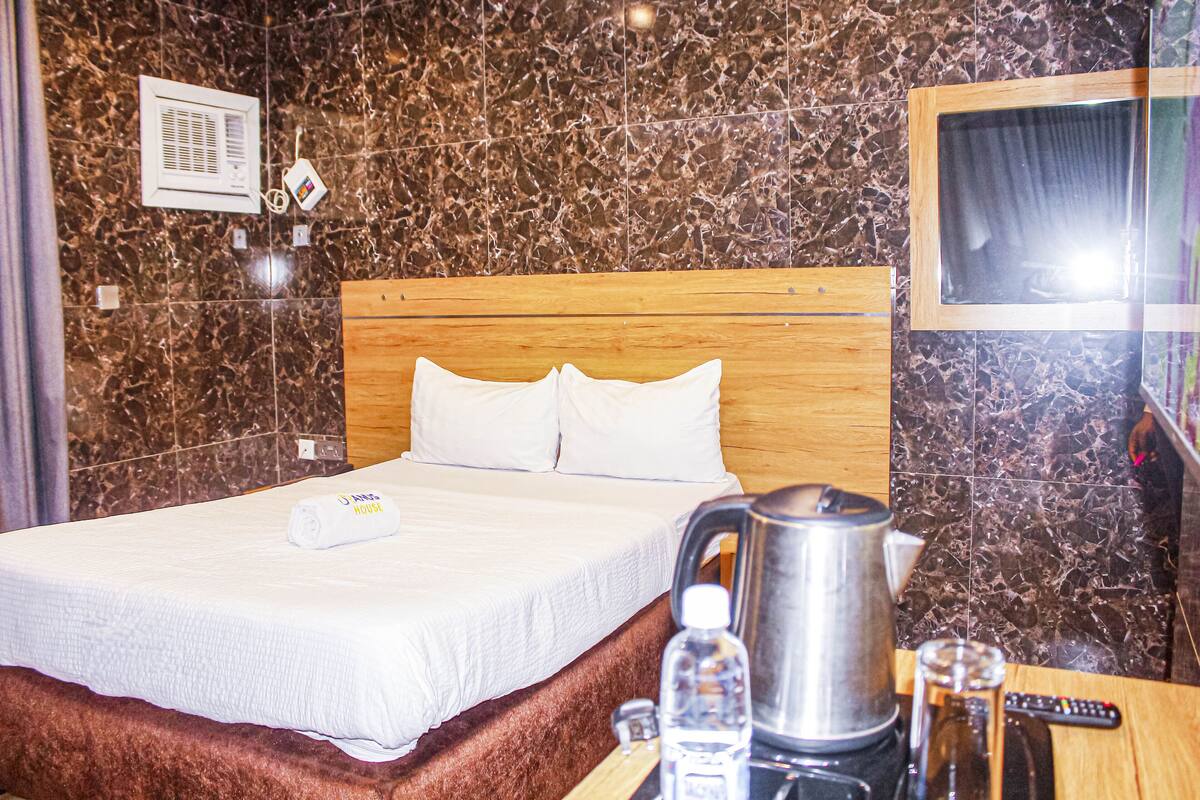 Ett hotellrum med en vägg som har ett marmormönster, en sänggavel i trä, en snyggt bäddad säng med vita sängkläder, en vattenkokare, ett glas vatten och en fjärrkontroll på sidobordet.