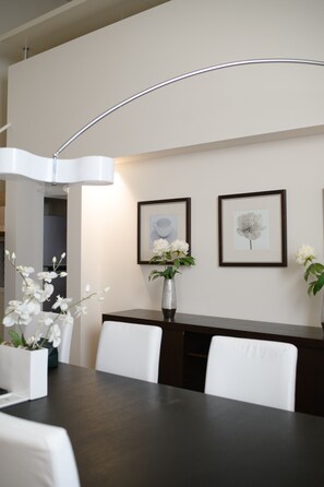 Een eethoek met een donker houten tafel, witte stoelen, een vaas met witte bloemen en een kunstwerk in een lijst aan de muur.