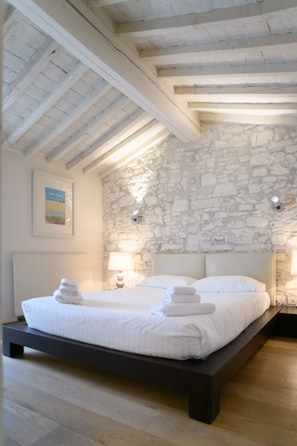 Een slaapkamer met een stenen muur, een houten plafond, een bed met witte lakens en twee bedlampjes.