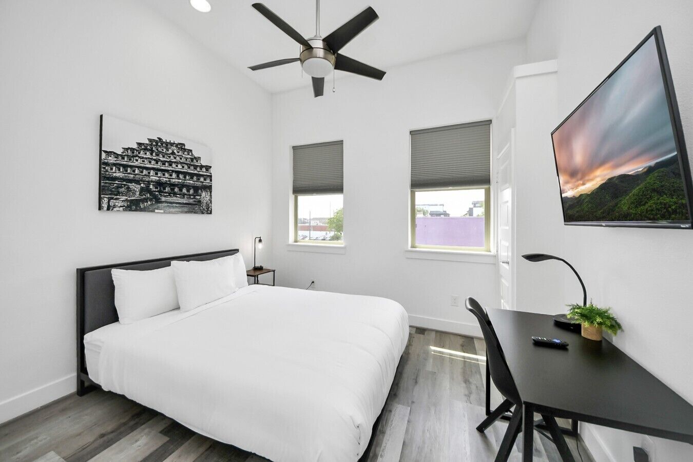 Habitación de hotel con cama, escritorio, ventilador de techo, dos ventanas y obras de arte en las paredes.