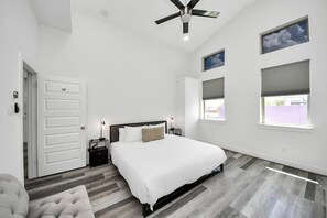 Un dormitorio con cama, ventilador de techo, mesita de noche y dos ventanas.