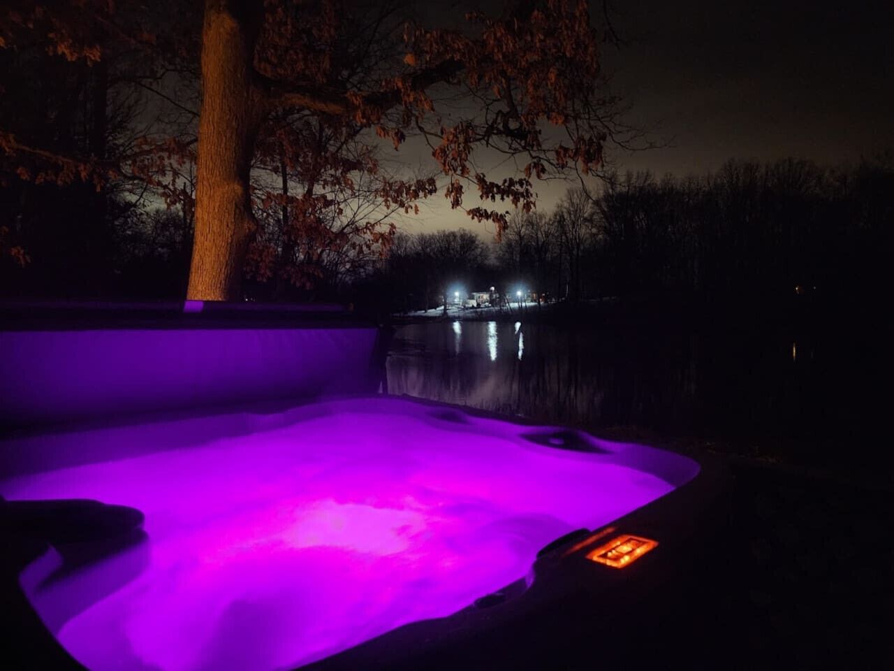 Un bain à remous doté d’un éclairage violet, entouré d’arbres et bordé par un plan d’eau.