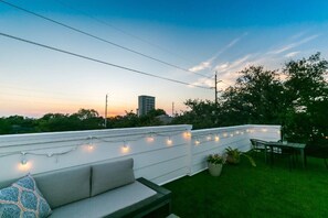 Eine Dachterrasse mit weißer Brüstung, Lichterketten, einer Couch und Stühlen.