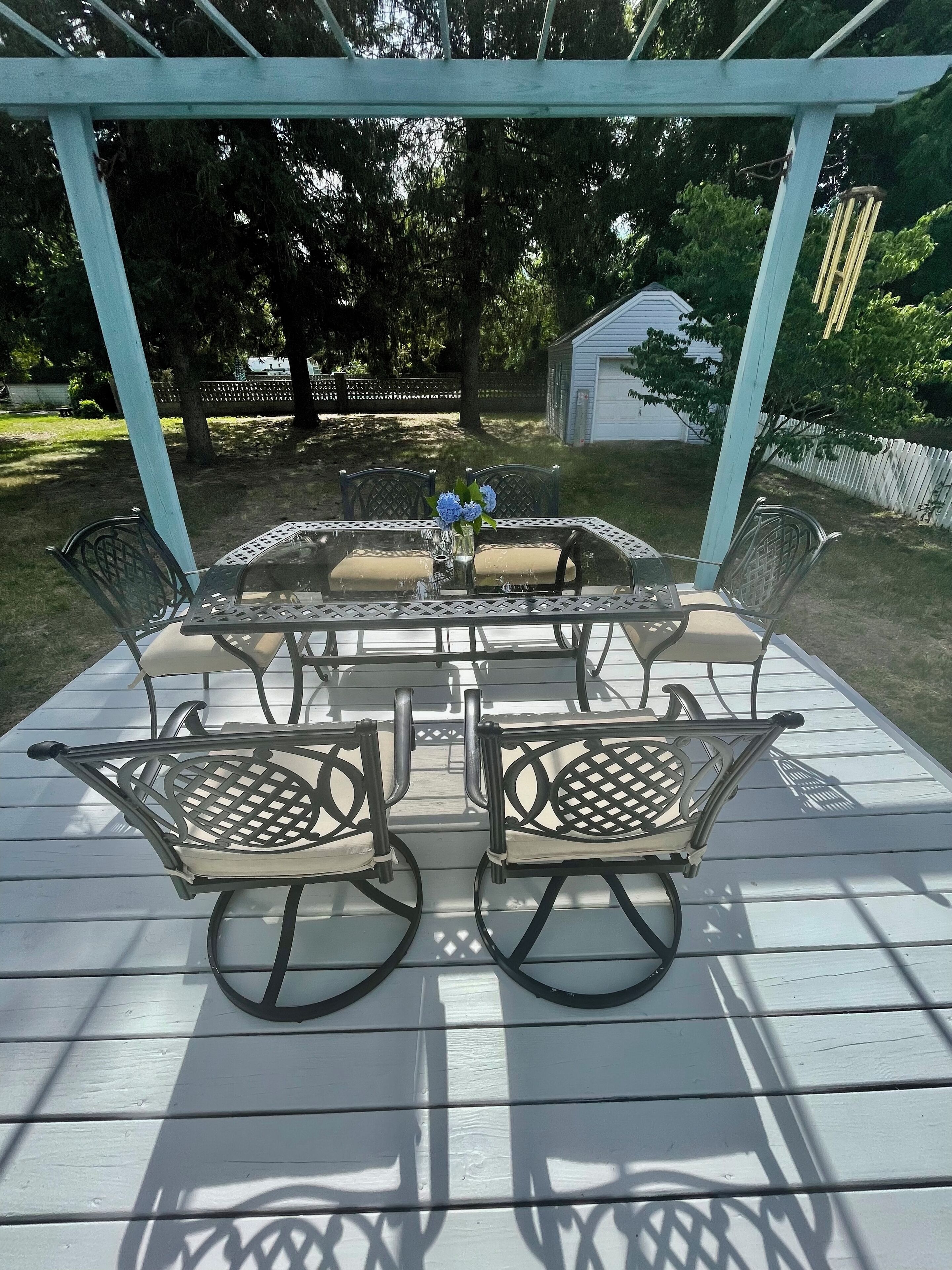 A covered patio with a glass-top table and four chairs, surrounded by a wooden deck and a white fence.