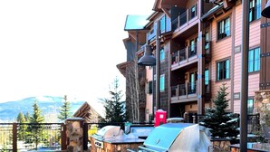 Un espace barbecue en pierre offrant une vue sur une chaîne de montagnes, un immeuble à plusieurs étages doté de balcons et une poubelle rouge.