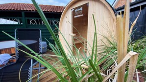 Une structure en bois portant l’inscription « HOME DELUXE », avec un toit recouvert de tuiles, entourée d’herbe haute et d’une chaise en métal.