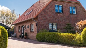 Une maison à deux étages, construite en briques, avec un toit recouvert de tuiles rouges, un petit porche doté de chaises et un jardin bien entretenu, agrémenté de haies et de plantes à fleurs.