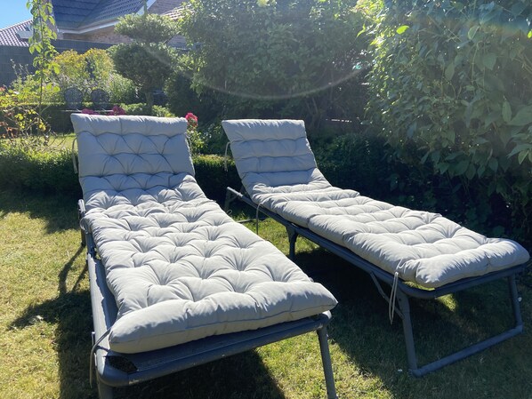 Two outdoor lounge chairs with cushioned seats and backs, positioned on a grassy lawn with a backdrop of dense foliage and a brick building.
