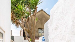 Un cortile stretto con pavimento in pietra, un albero, un muro di pietra, un ombrellone a righe blu e un tavolo e una sedia in metallo.