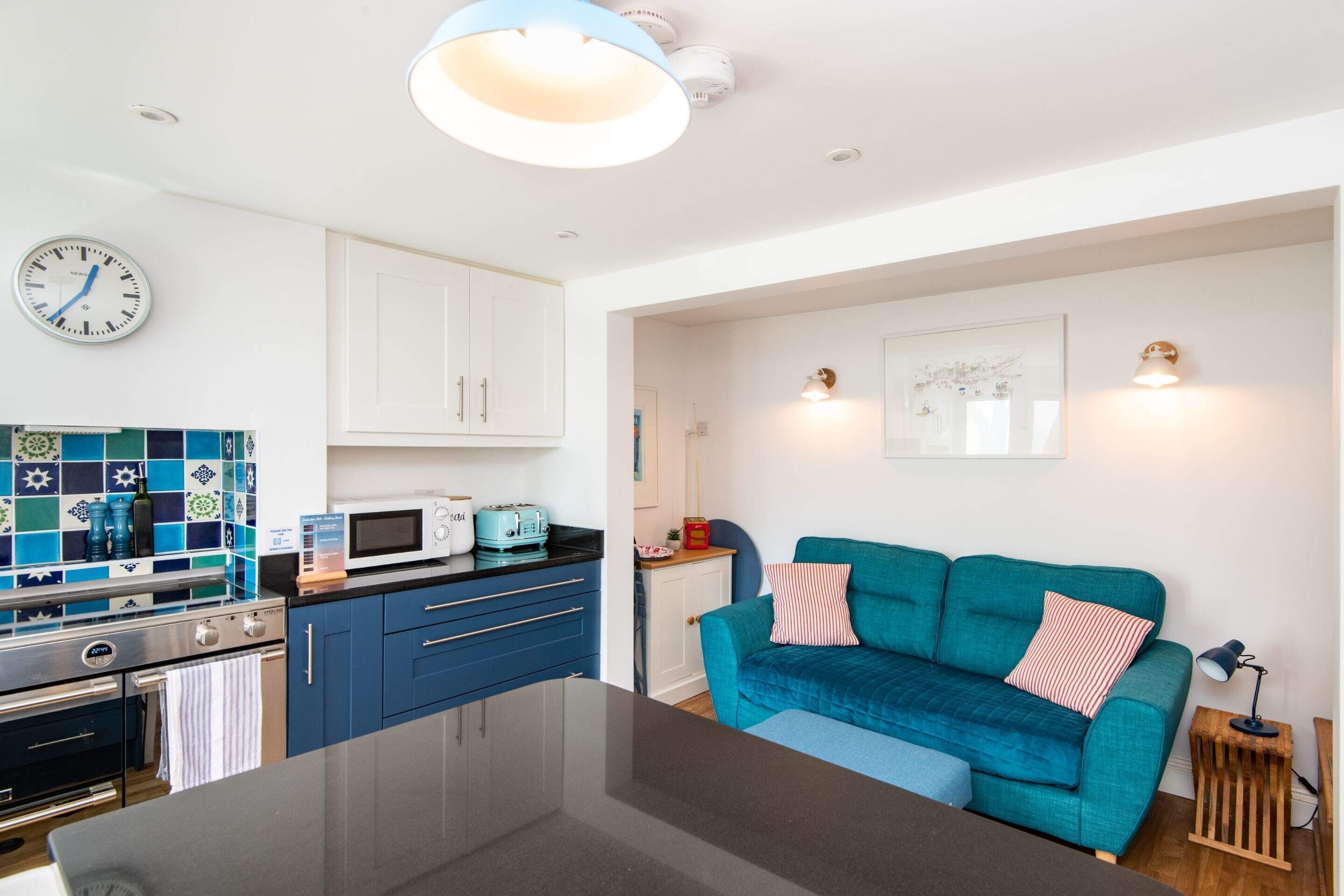 A modern kitchen with white cabinets, a blue countertop, and a microwave. There is a teal sofa with a striped pillow and a small wooden side table with a lamp.