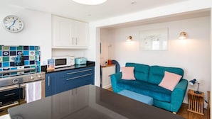 Una cucina moderna con mobili bianchi, un piano di lavoro blu e un forno a microonde. C'è un divano verde acqua con un cuscino a righe e un piccolo tavolino in legno con una lampada.