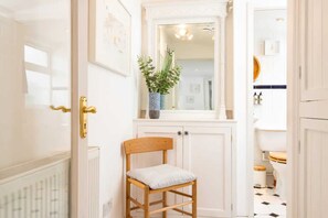 A narrow hallway with a wooden chair, a white radiator, and a window with a plant. - Quirky 1 Bedroom Apartment in Kemptown (Brighton)