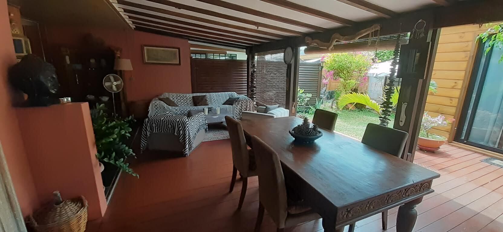 Een overdekt terras met een houten tafel en stoelen, een zithoek met kussens, en uitzicht op een tuin.