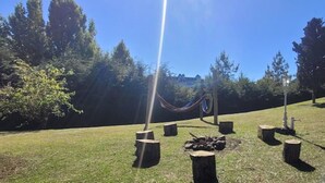 Ein grünes Areal im Freien mit einer Hängematte, Holzpfosten und einer Feuerstelle.