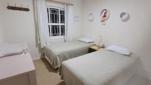 Ein Zimmer mit zwei Betten, einem Fenster mit weißen Vorhängen, einem kleinen Tisch mit einer Lampe sowie Wanddekorationen, darunter ein Vogel und eine Platte.