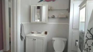 A small, white bathroom with a toilet, a sink with a mirror above it, and a shelf with toiletries.