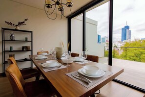 A dining area with a table set for four, a modern chandelier, and a city view through large windows. - Capitalia | Del Valle 2BR · Terrace & Parking (Ciudad de México)