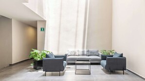 A modern lounge area with a grey sofa, two armchairs, and a coffee table. There are potted plants and a visible emergency exit sign.