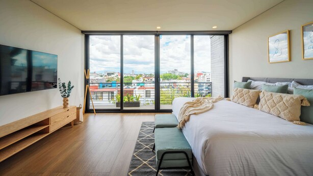 A modern bedroom with a large bed, a TV, and a balcony with a city view.
