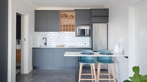 A modern kitchen with grey cabinets, a white countertop, a microwave, and a sink. There are two blue bar stools and a white kettle on the counter. - HoneyBee Cottage (Yzerfontein)