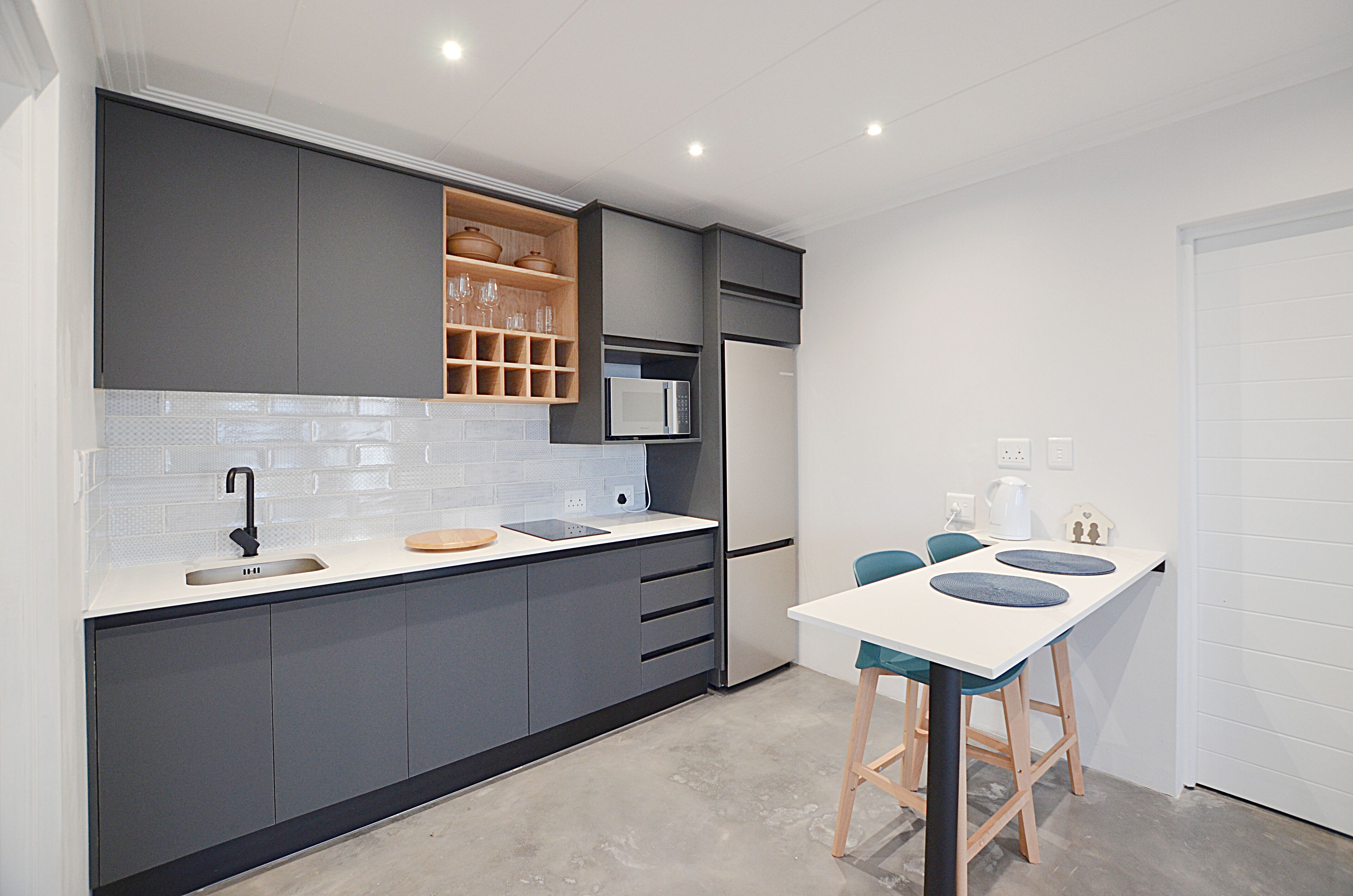 A modern kitchen with grey cabinets, a white countertop, and a built-in microwave. There is a small dining area with a white table and blue chairs.