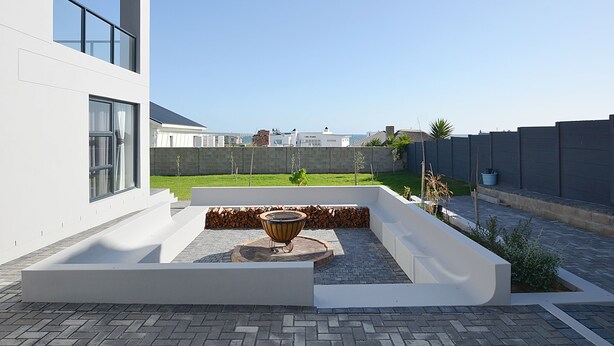 A modern outdoor patio with a fire pit, a white wall, and a view of a residential area. - HoneyBee Cottage (Yzerfontein)