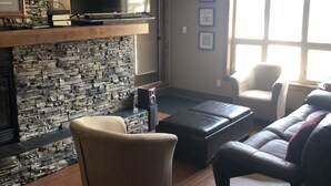 A living room with a stone fireplace, a flat-screen TV, a ceiling fan, a sofa, and a window with natural light.