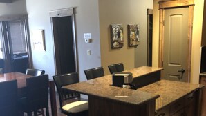 A kitchen with a central island, bar stools, a dining table, and chairs. There is a clock on the wall and a door with a handle.