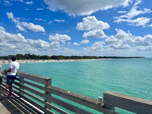 A wooden pier with a person standing at the edge, overlooking a beach with clear turquoise water and a line of trees in the background. - New Luxurious Coastal Resort 3-Beedroom 2 Bath Private Pool
(Venice)