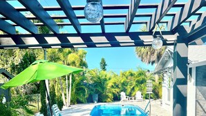 Un espacio exterior techado con piscina, sillas de descanso y una pérgola con una lámpara colgante.