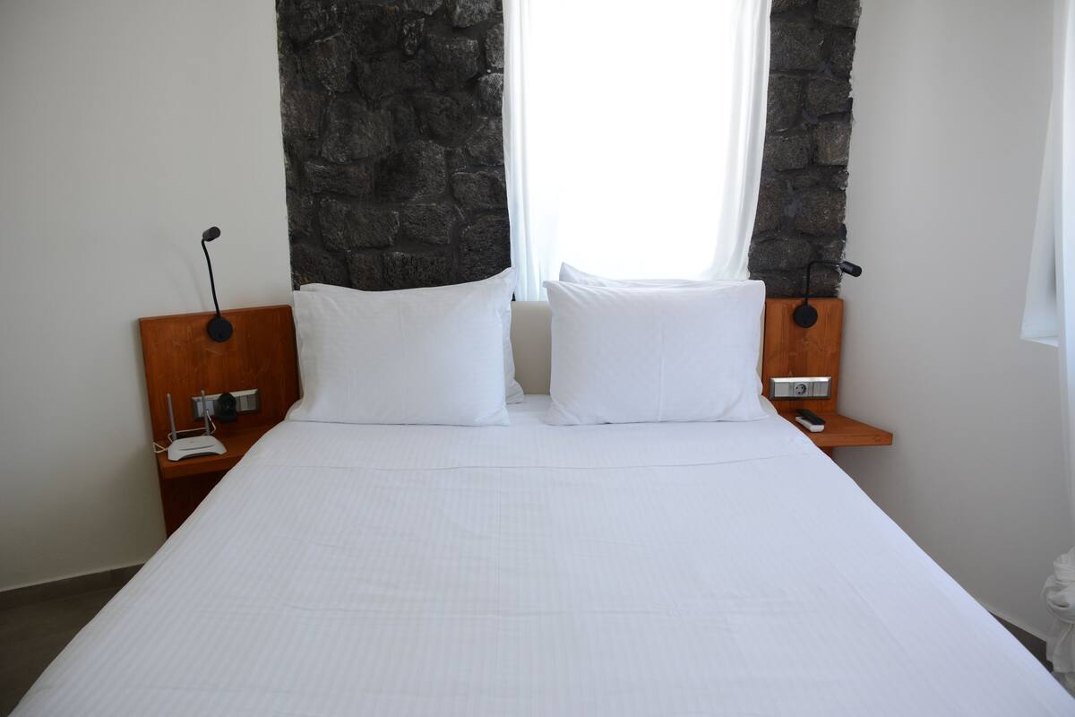 Una cama bien hecha con sábanas blancas, cabecera de madera y pared decorativa de piedra.