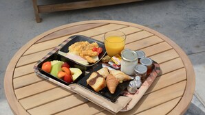 A round wooden table with a meal tray containing a sandwich, croissant, fruit, and juice. - Noir Santorini (Santorini)