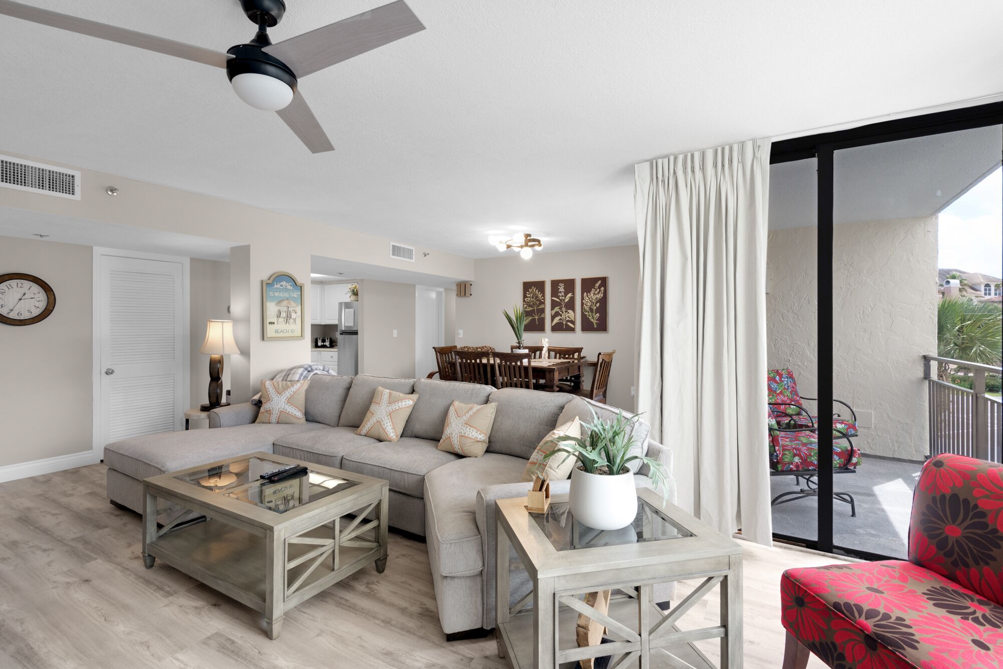 A living room with a sofa, coffee table, and a red chair. There is a dining area with a table and chairs, and a balcony with a chair.