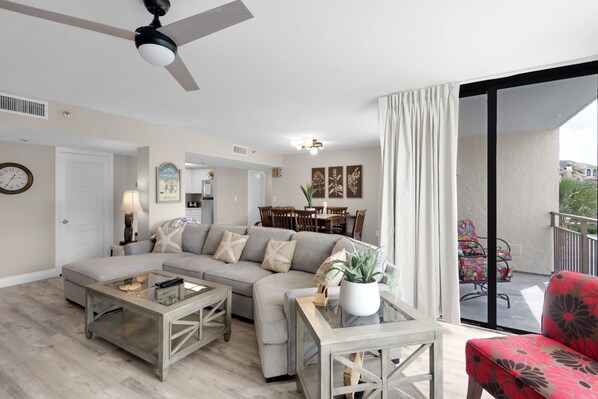 Sala de estar com sofá, mesa de centro e uma cadeira vermelha. Há uma área de jantar com mesa e cadeiras, e uma varanda com uma cadeira.