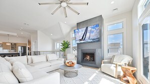 A modern living room with a fireplace, a large TV mounted above it, and a view of the outdoors through large windows. - BEACHFRONT Multi-Family Home, 3 levels with decks! (Brigantine)