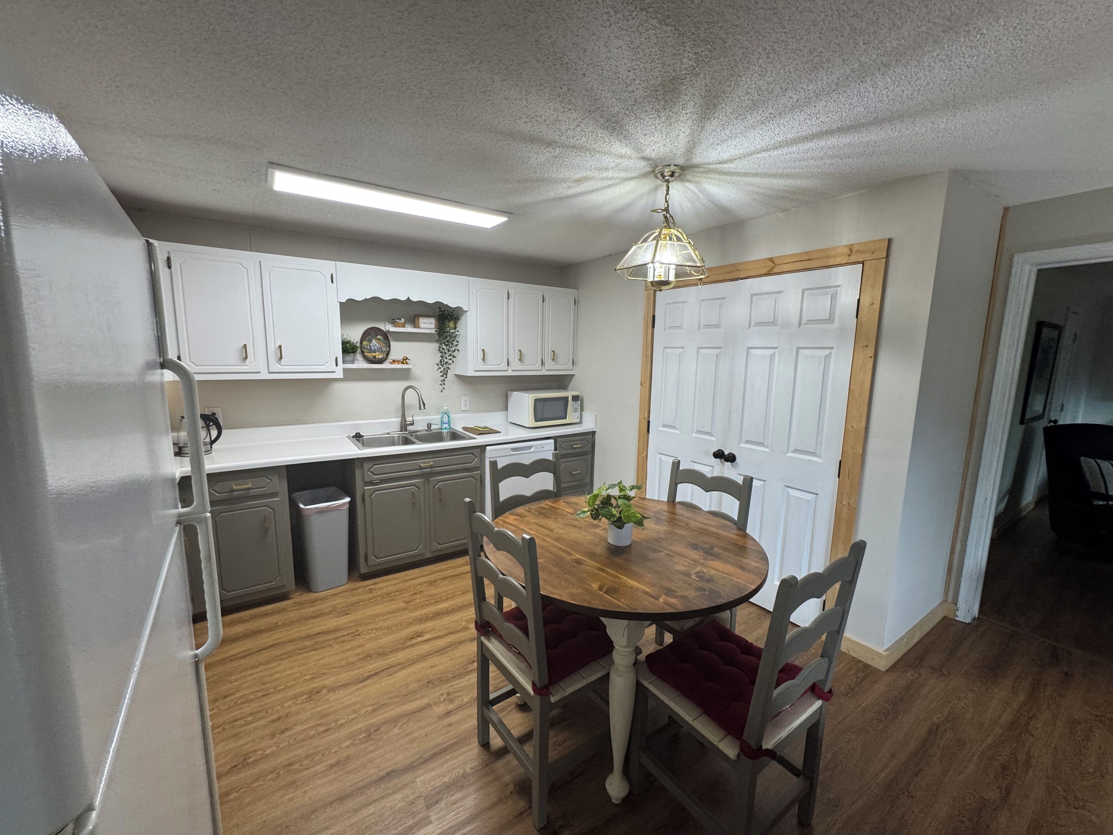 A compact kitchen with white cabinets, a wooden dining table with chairs, a microwave, and a ceiling fan.