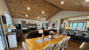 Un salón amplio con techo de madera, chimenea, una mesa de comedor con sillas y una zona de cocina con refrigerador y pileta.
