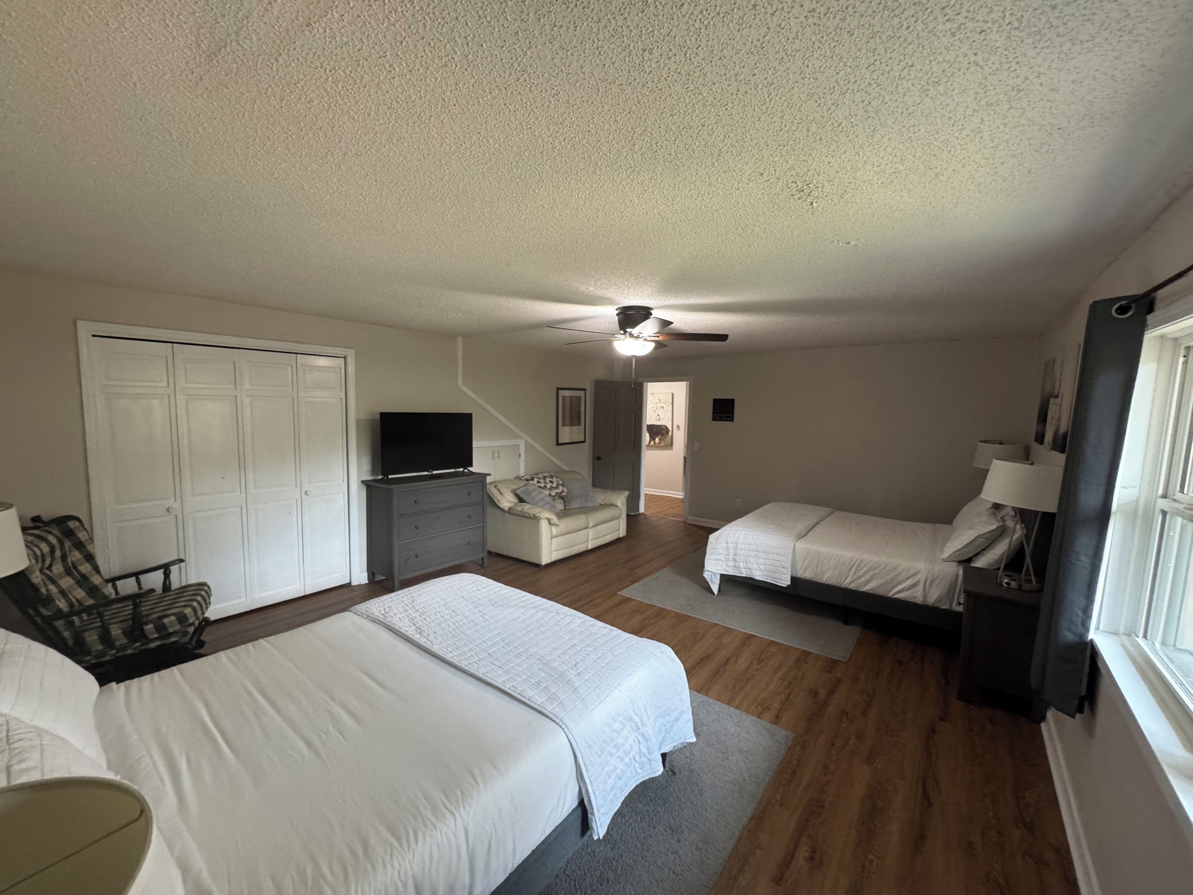 Une chambre avec deux lits, un bureau, une chaise, un ventilateur de plafond et une porte donnant sur une autre pièce.