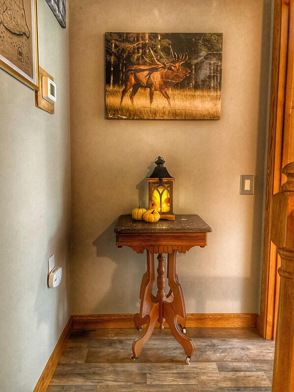 Un couloir avec une table en bois sur laquelle sont posés un lanternon et des citrouilles, un tableau représentant un cerf et un plancher en bois.