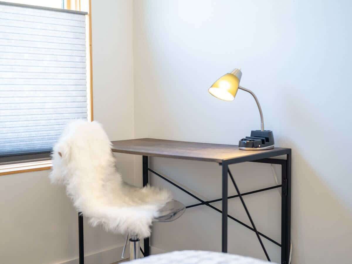 A small desk with a lamp, a fluffy chair, and a window with blinds.