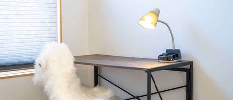 A small desk with a lamp, a fluffy chair, and a window with blinds.