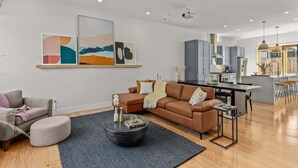 Een moderne woonkamer met een bruine bank, een ronde salontafel en een grijs vloerkleed. Achtergrond toont een keuken met grijze kastjes en een bar.