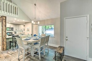Une cuisine spacieuse avec des meubles blancs, un coin repas comprenant une table en bois et des chaises blanches, et une vue sur l’extérieur grâce à une grande fenêtre.