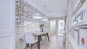 Un comedor con piso de madera en diseño espiga, paredes blancas y obras de arte enmarcadas. Dos lámparas colgantes iluminan la mesa.