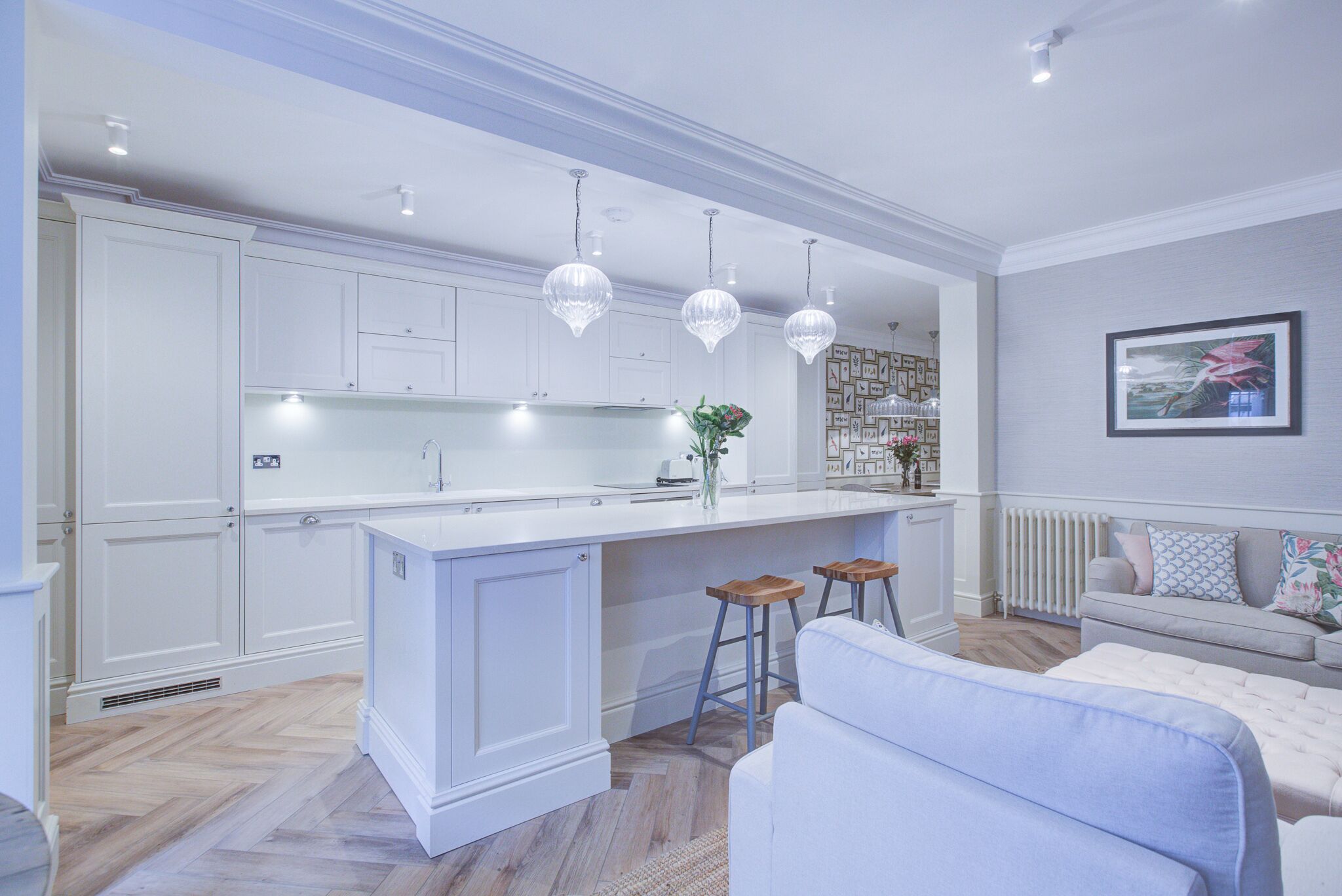 Una cocina moderna con armarios blancos, una isla central, luces colgantes y una zona de estar con sofá y taburetes.
