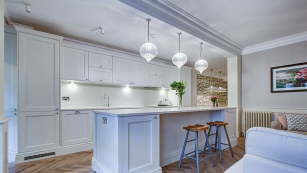 Cocina moderna con armarios blancos, una isla central, taburetes de bar, luces colgantes y un radiador.