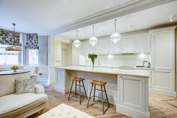 A modern kitchen with white cabinetry, a central island with stools, pendant lighting, and a view of the adjacent living area. - Stylish 2 bedroom apartment with private courtyard garden (London)