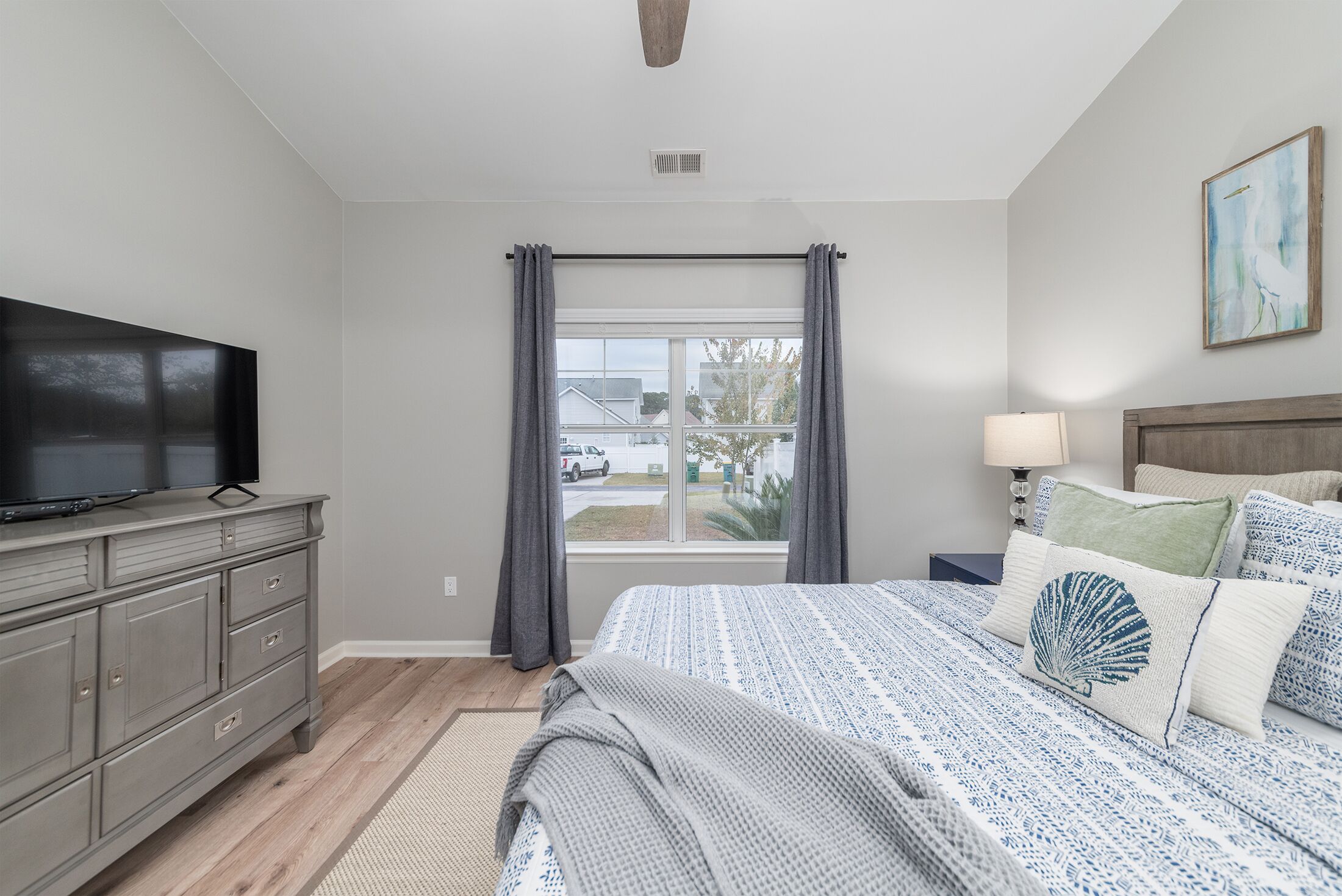 A bedroom with a bed, a dresser, a TV, a window with curtains, a lamp, and a painting on the wall.